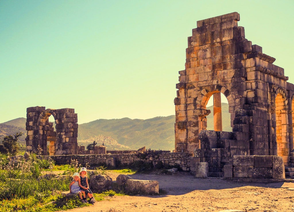2 small girls sat wearing sun hats, sat in the sun in front of Roman remains at Volubilis, Morocco