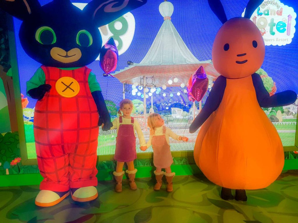 2 small girls stood between and holding hands with 2 large costumed characters from the Kid's TV show, 'Bing'