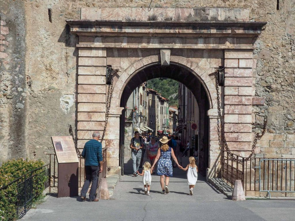 Mother holding hands with 2 daughters, walking through a large, stone, town entrance gate