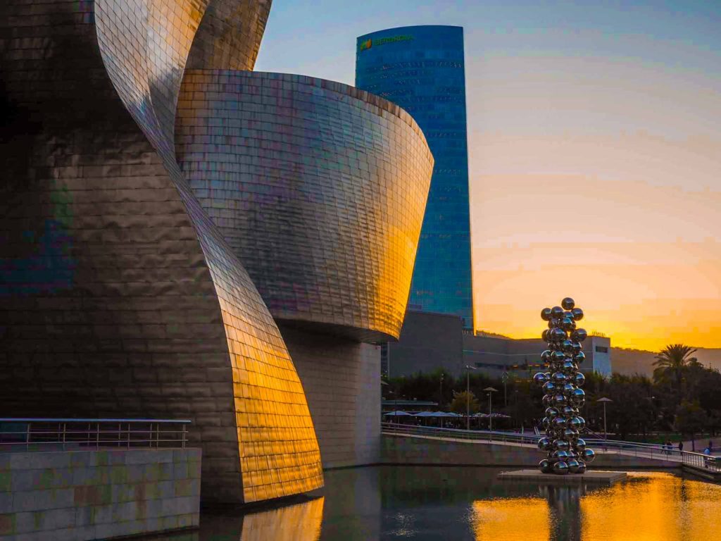 Orange sunset light reflecting off the shiny, curved shapes that form the building exterior of the Guggenheim Museum, Bilbao