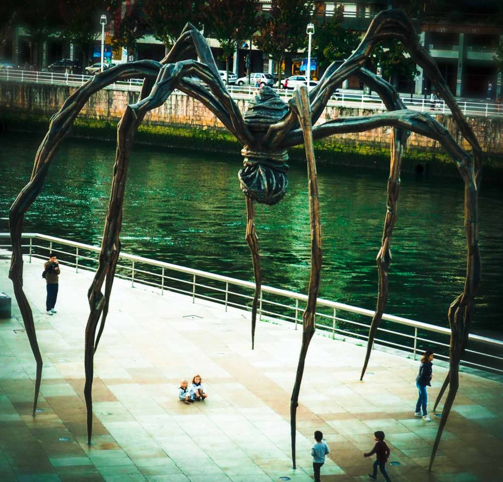 2 small girls sat under a giant metal spider sculpture, outside the Guggenheim Museum, Bilbao
