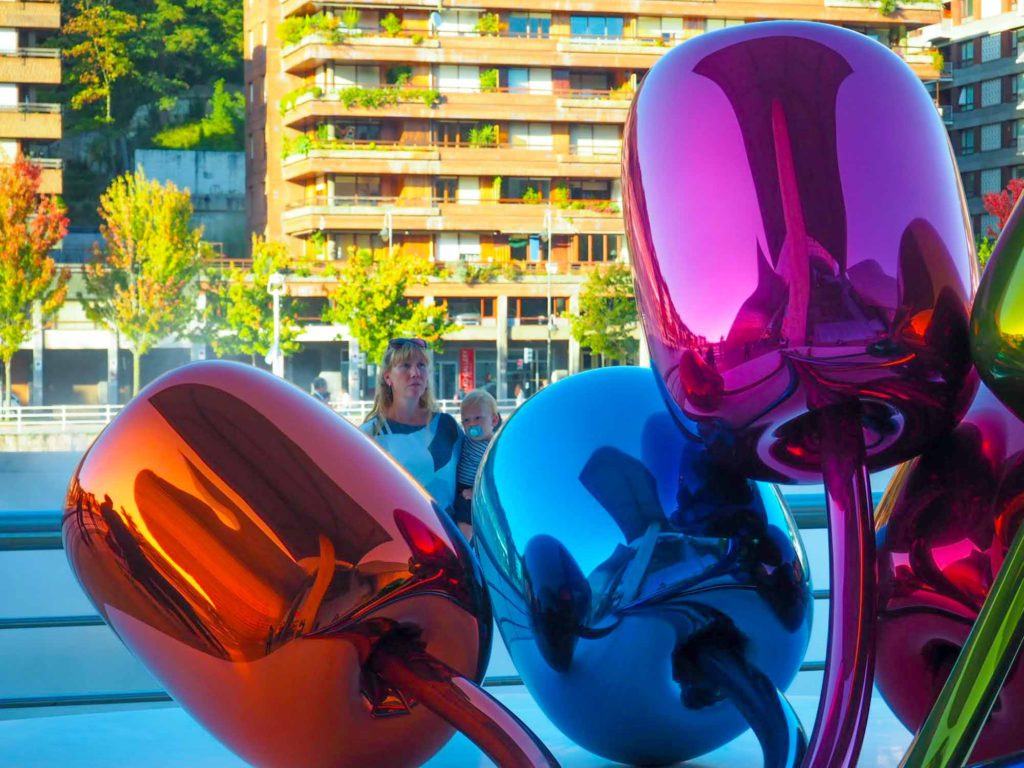 Mother holding a baby boy while looking at a large, colourful, metal tulip flower sculpture, outside the Guggenheim Museum, Bilbao