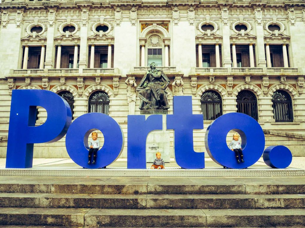3 small children sat inside the letters of a sculpture of giant blue lettering, spelling 'Porto'