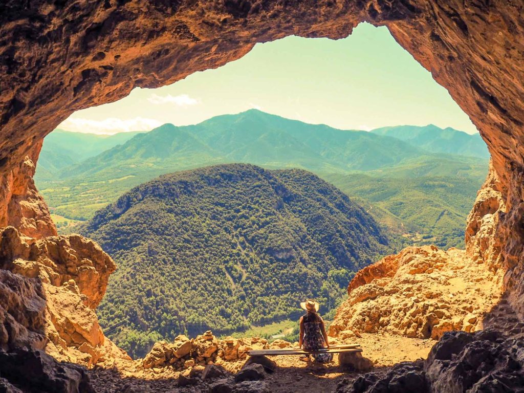 Lady sat on a wooden bench at the entrance to a cave high in the Pyranees mountains, with mountain views behind