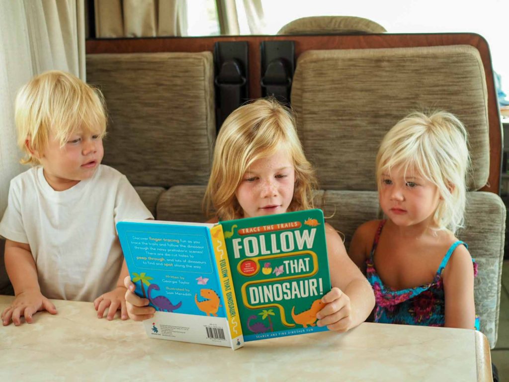 3 small children sat at a table in a motorhome, reading the book 'Follow that Dinosaur!'