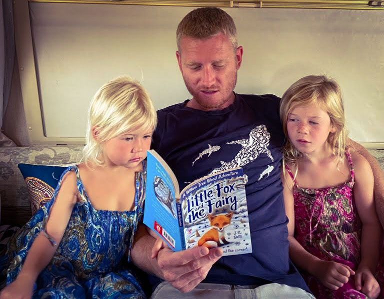 Father reading a story book to 2 young girls, sat inside a motorhome