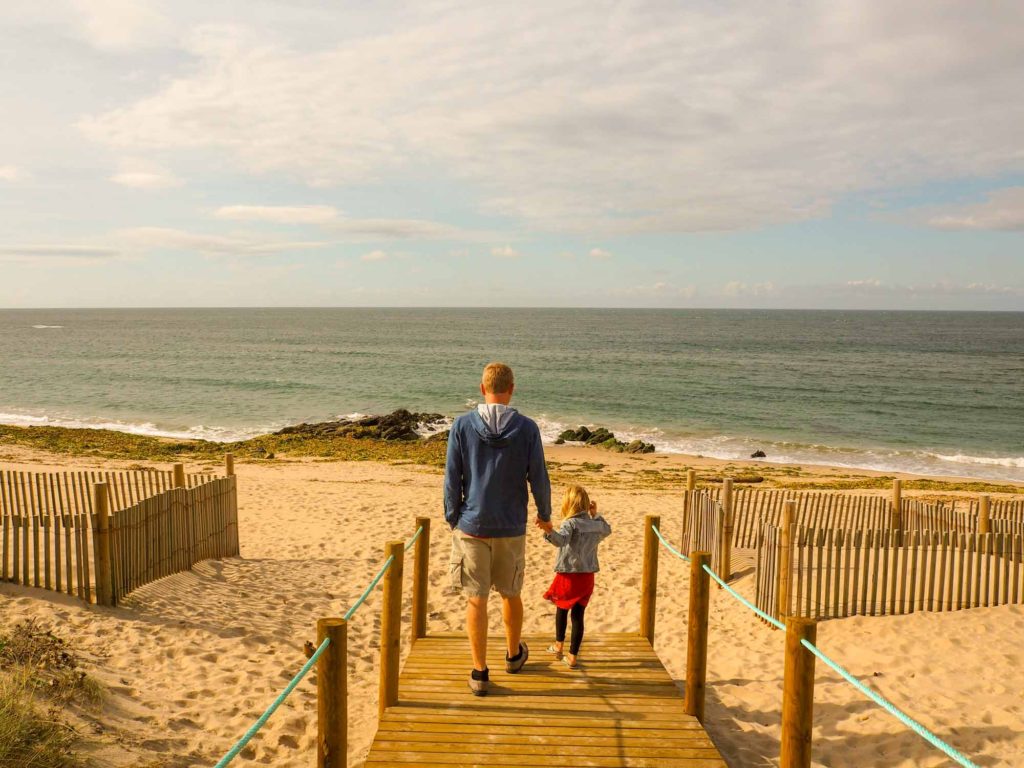 Father and daughter holding hands and walking onto a sandy beach from a boardwalk