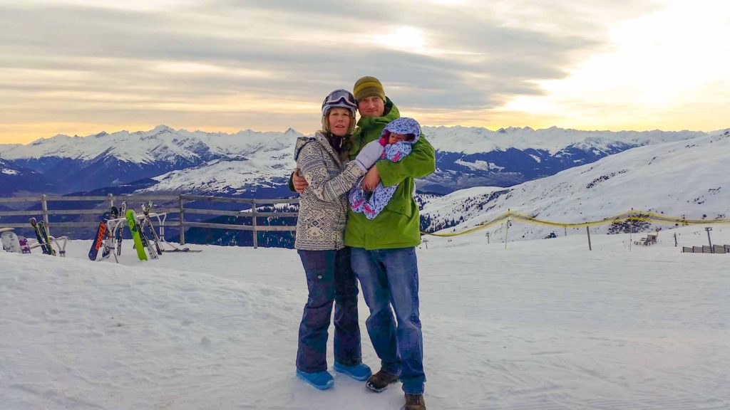 Man and a woman wearing ski clothes and holding a baby, with snowy mountain scenery behind