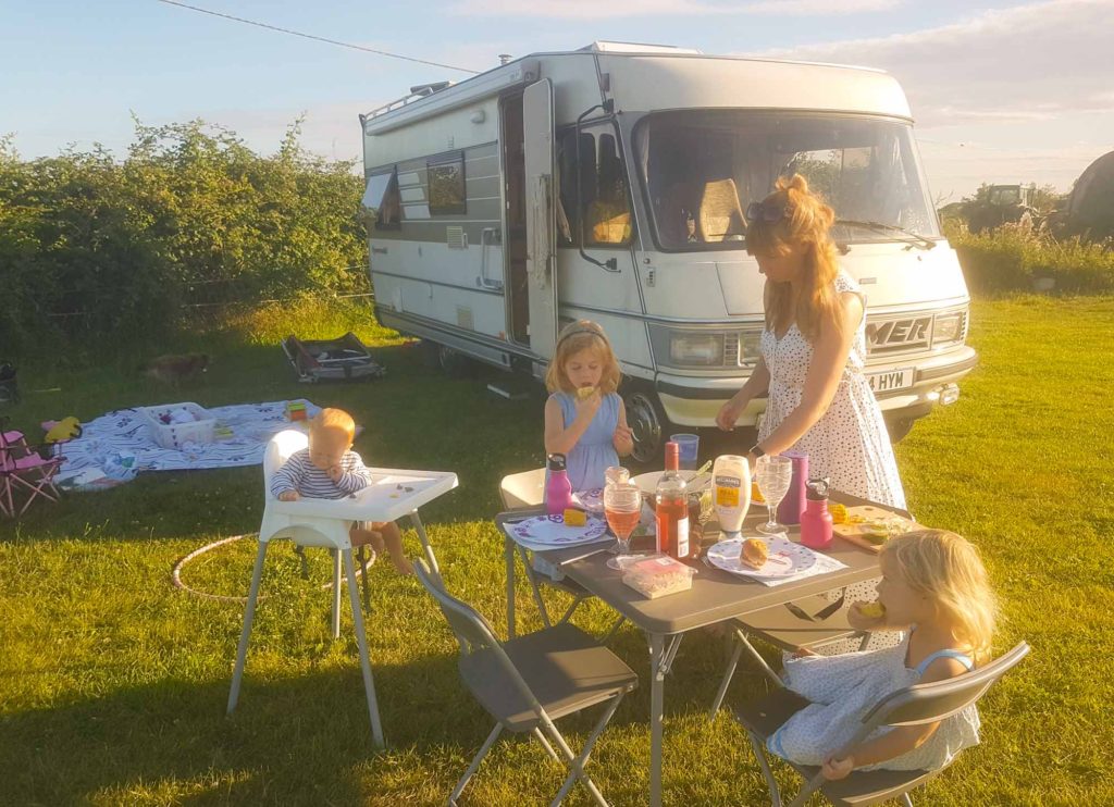 Family eating dinner outside at a camp table, next to a classic Hymer motorhome