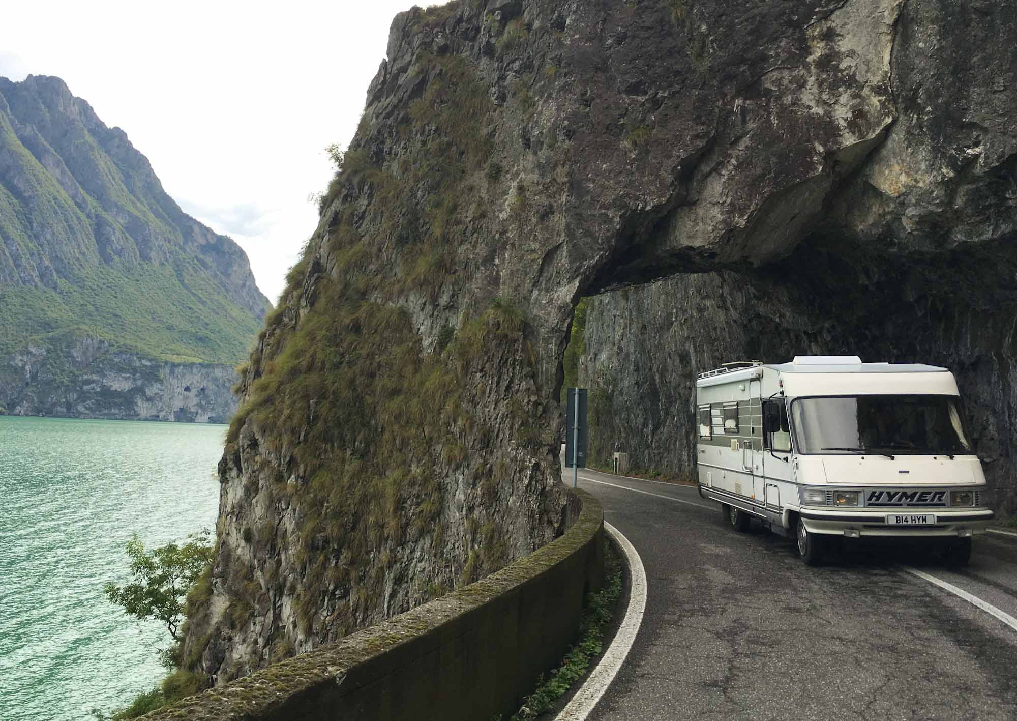 Classic Hymer motorhome driving through a natural rock road tunnel, by the side of a lake