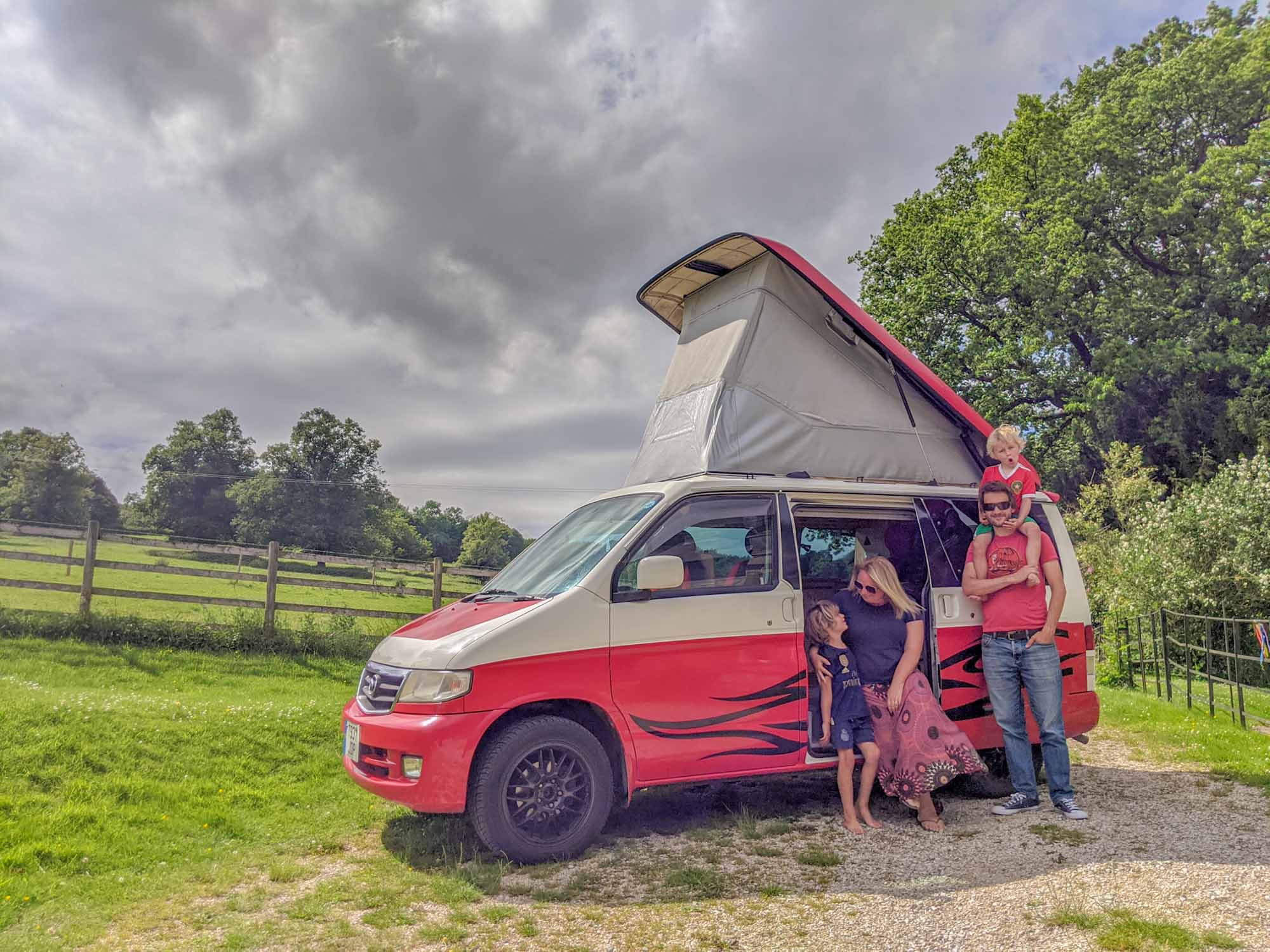 Family gathered around their red, Mazda Bongo camper van in the countryside, with roof raised for camping