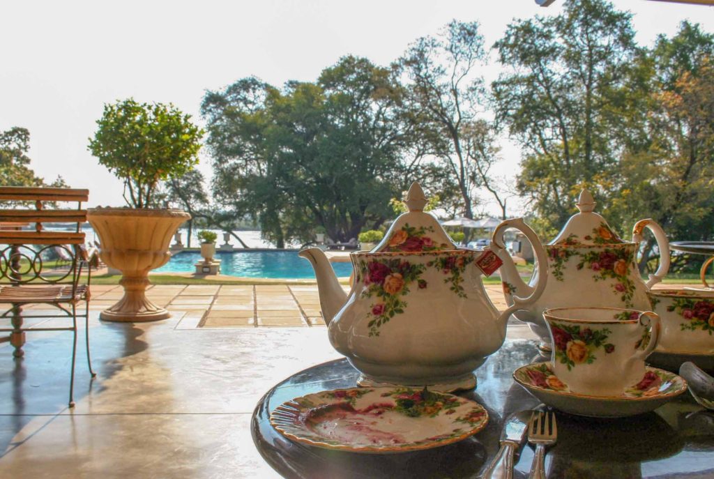 Decorative bone china teapot and tea cups arranged on a table, with view out to a swimming pool and Zambezi River behind