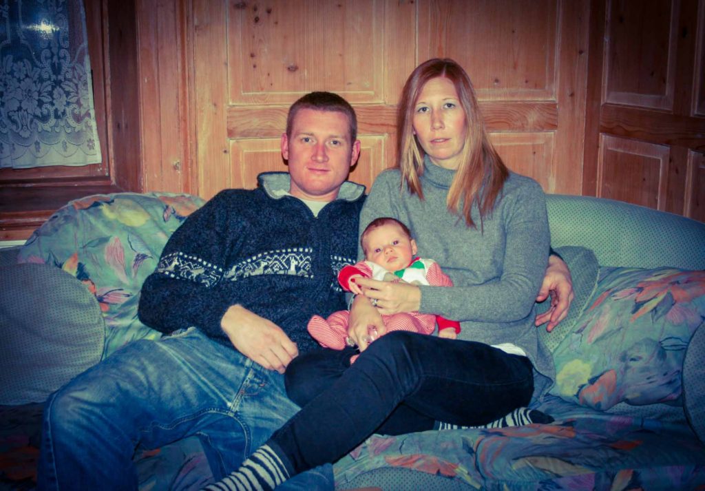 Mother and father sat holding a baby, in a ski chalet, with wooden wall panelling behind