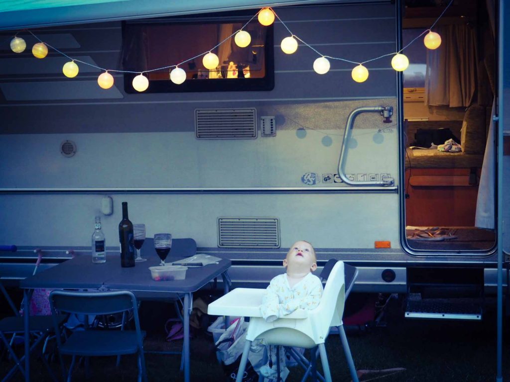 Small child sat in a highchair outside a motorhome, looking up at hanging string lights