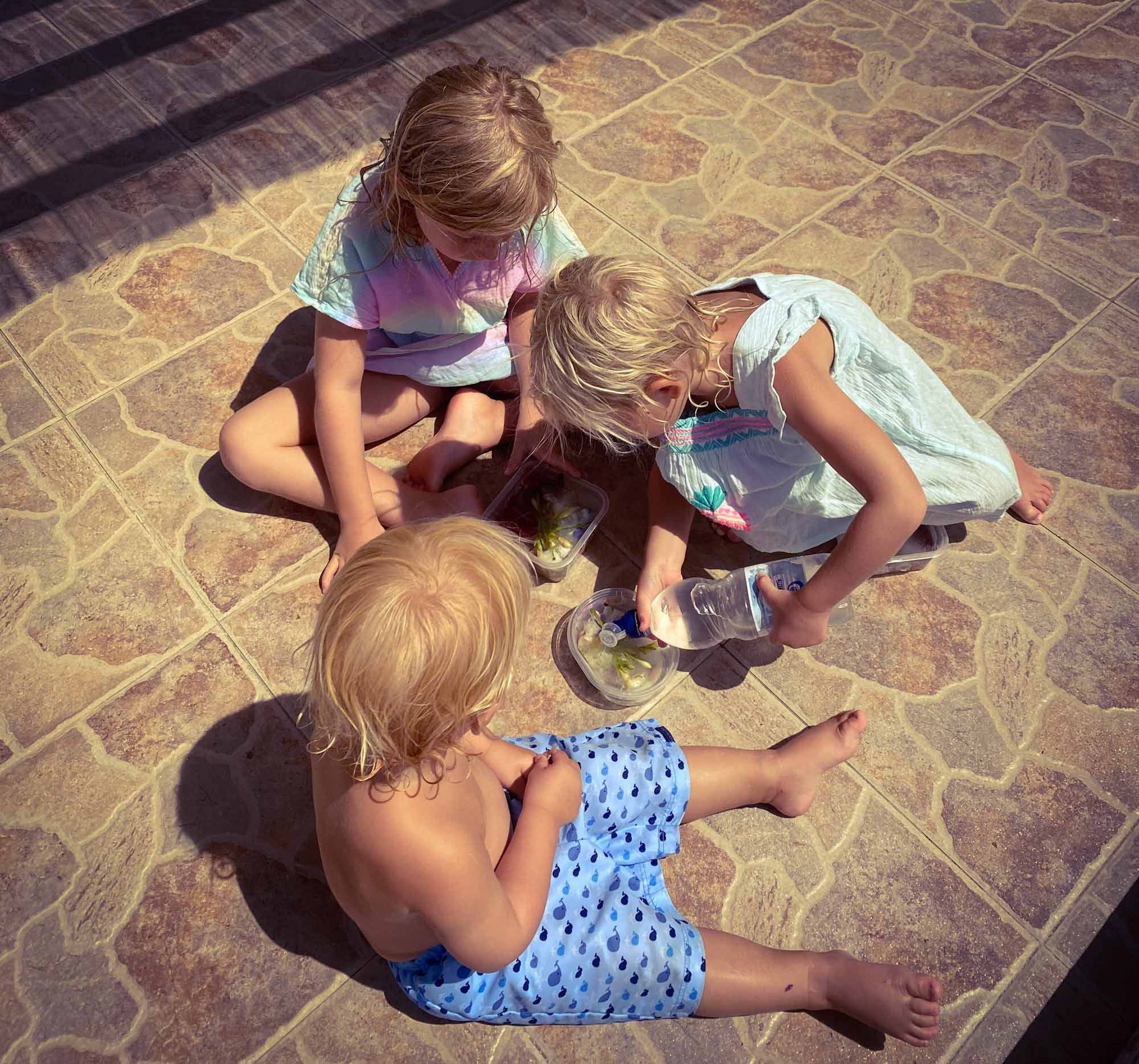 3 small children sat on the floor outside, as one pours water from a bottle into a plastic tub