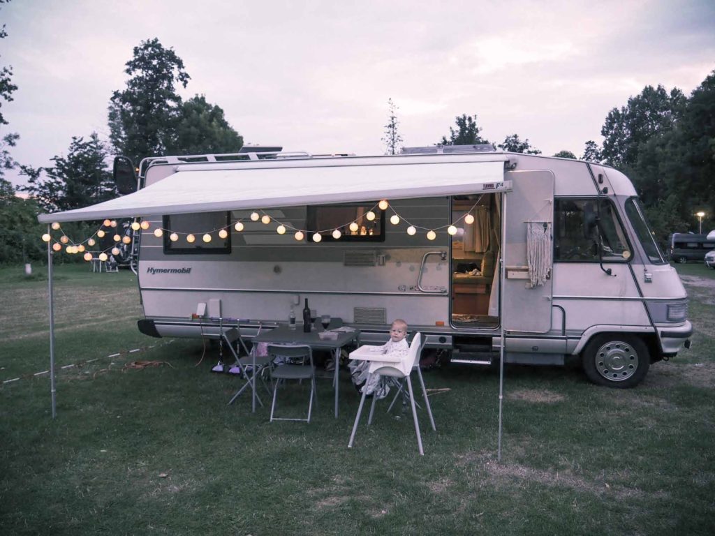 A small child sat outside old motorhome, with string lights hanging from the rollout awning
