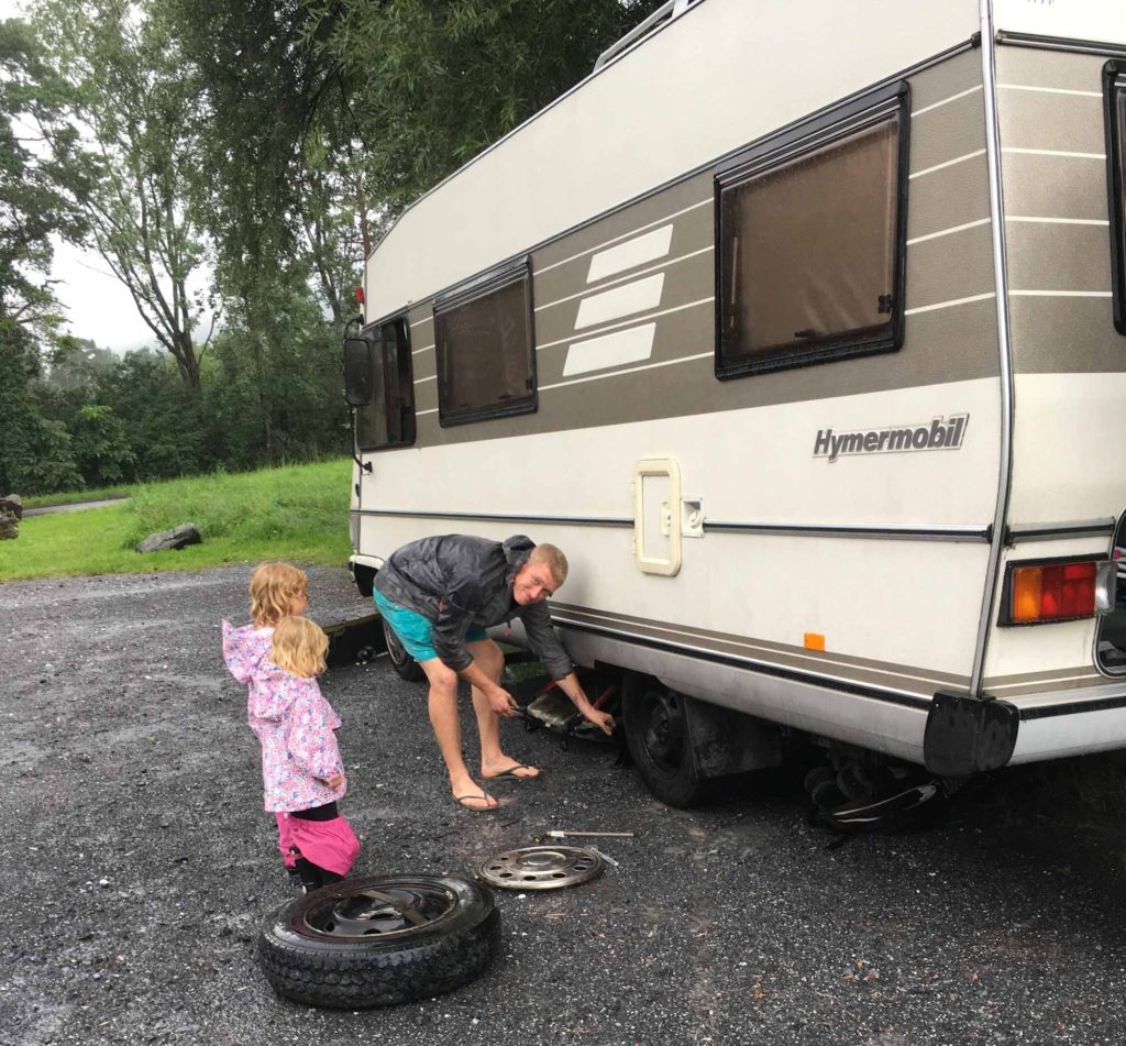 Man changing a tyre on an old motorhome, with 2 small girls watching