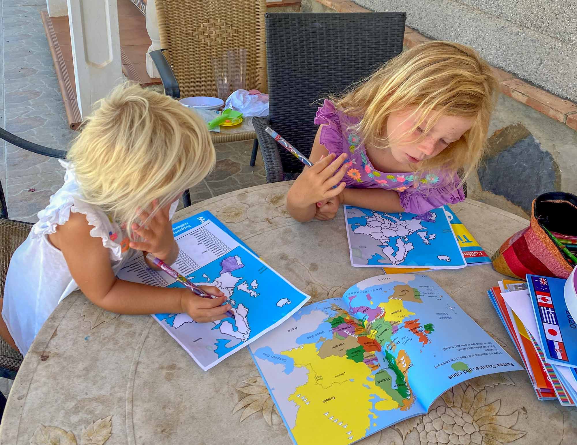 2 girls sat at a table colouring in a map of Europe, by referring to an atlas book