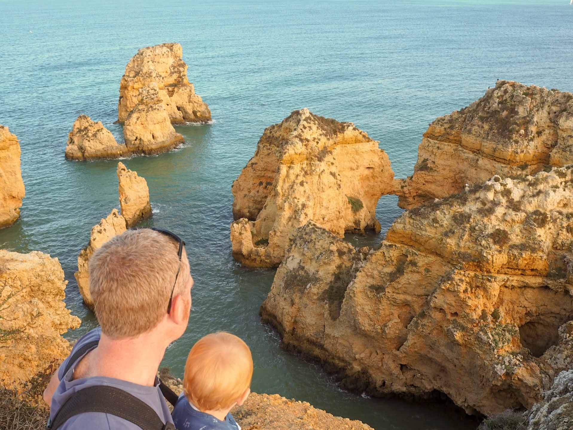 Father holding a small child while peering over a cliff edge down to the sea below, with eroded rock formations