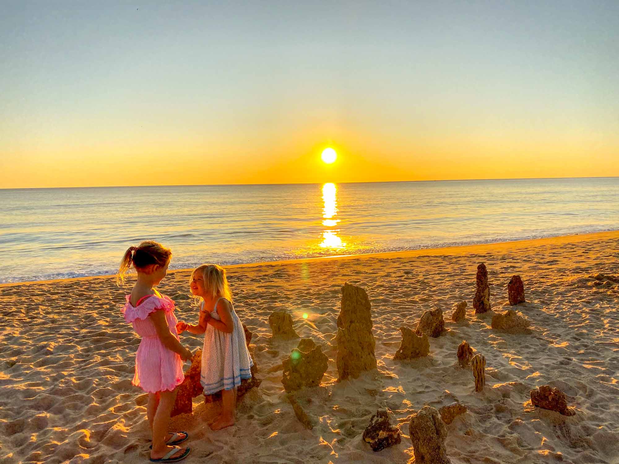 2 small girls stood on a sandy beach with the sun setting behind