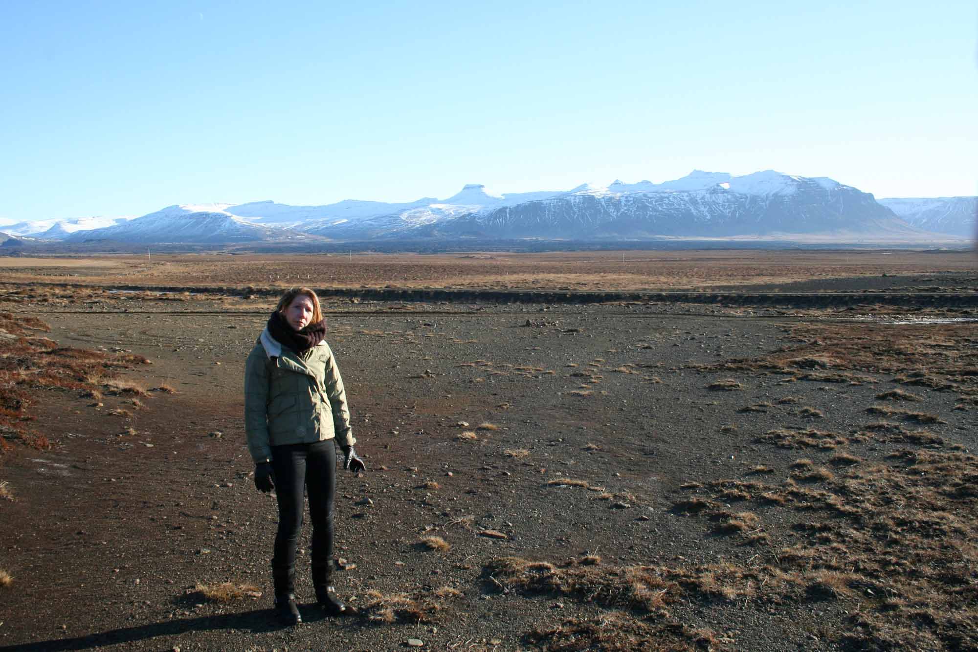 Lady stood in a barren, black sand landscape in Iceland, with snowy mountains behind