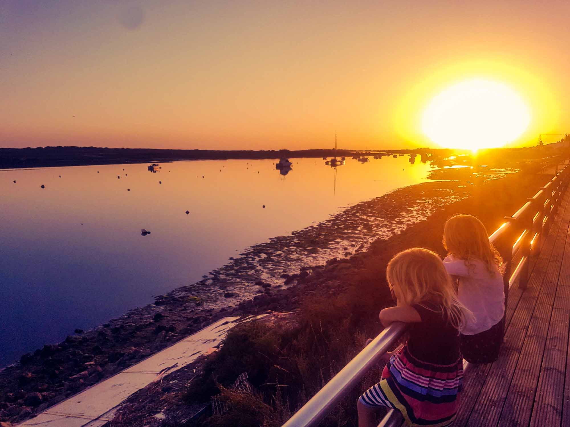 2 girls sat on metal rails of a fence, looking out over water with boats on, at sunset