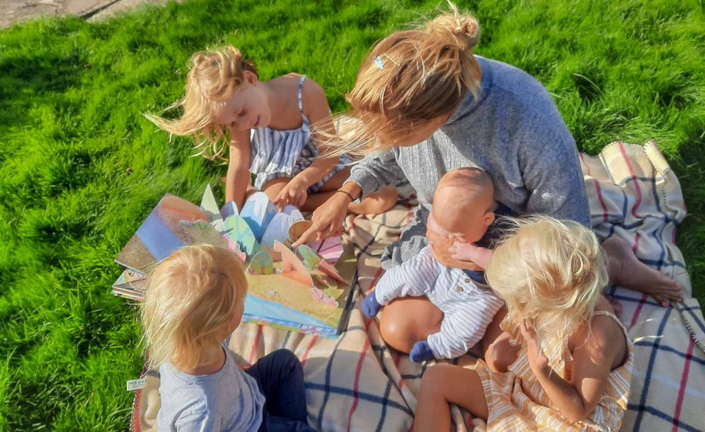 Family with small children sat outside on a rug while looking at a pop-up book