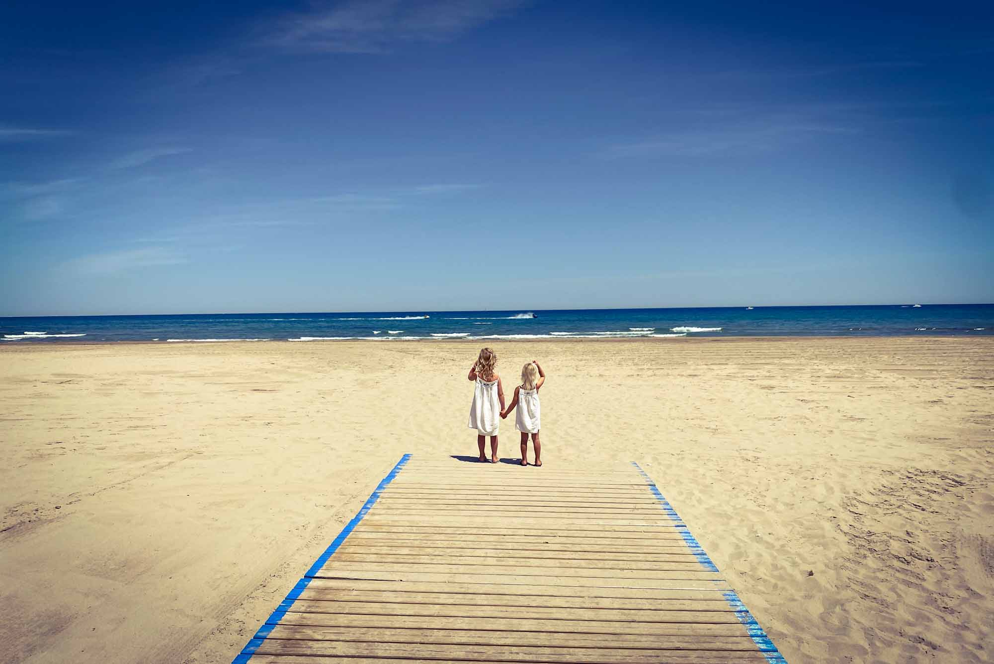 2 young girls holding hands at the entrance to a large and empty, sandy beach