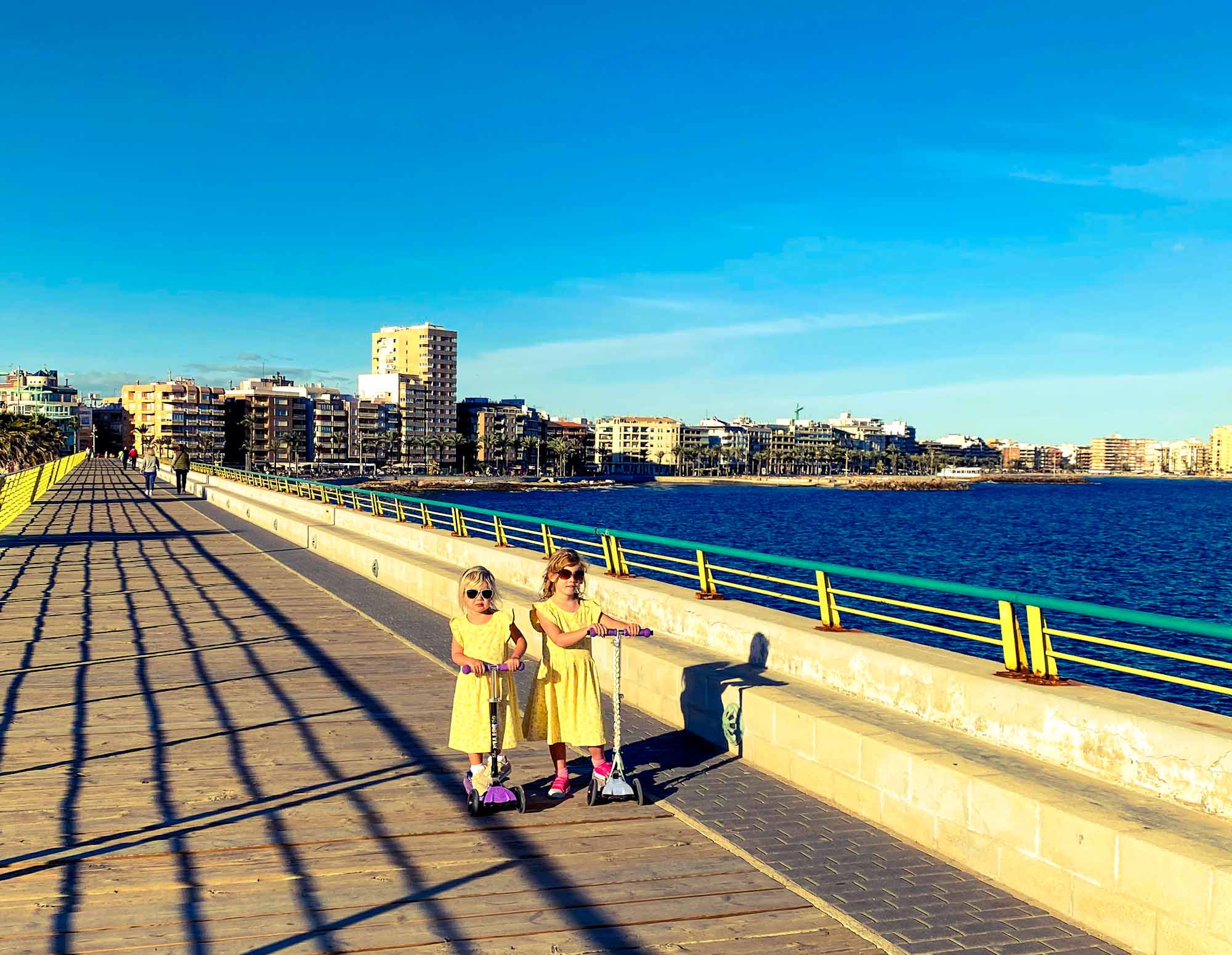 2 girls riding scooters along a seafront boardwalk