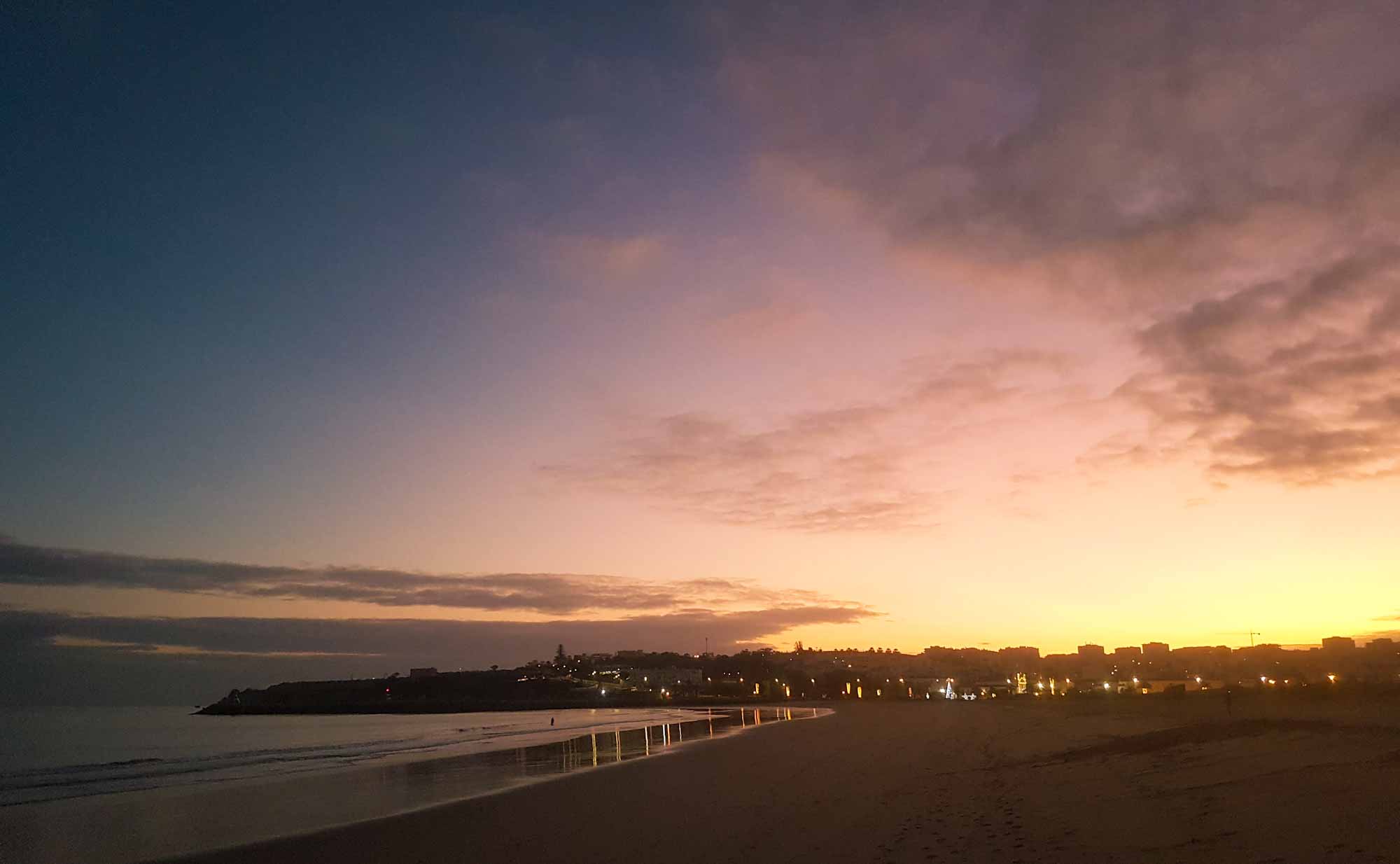 View of a long sandy beach at sunset, with the lights of a town behind