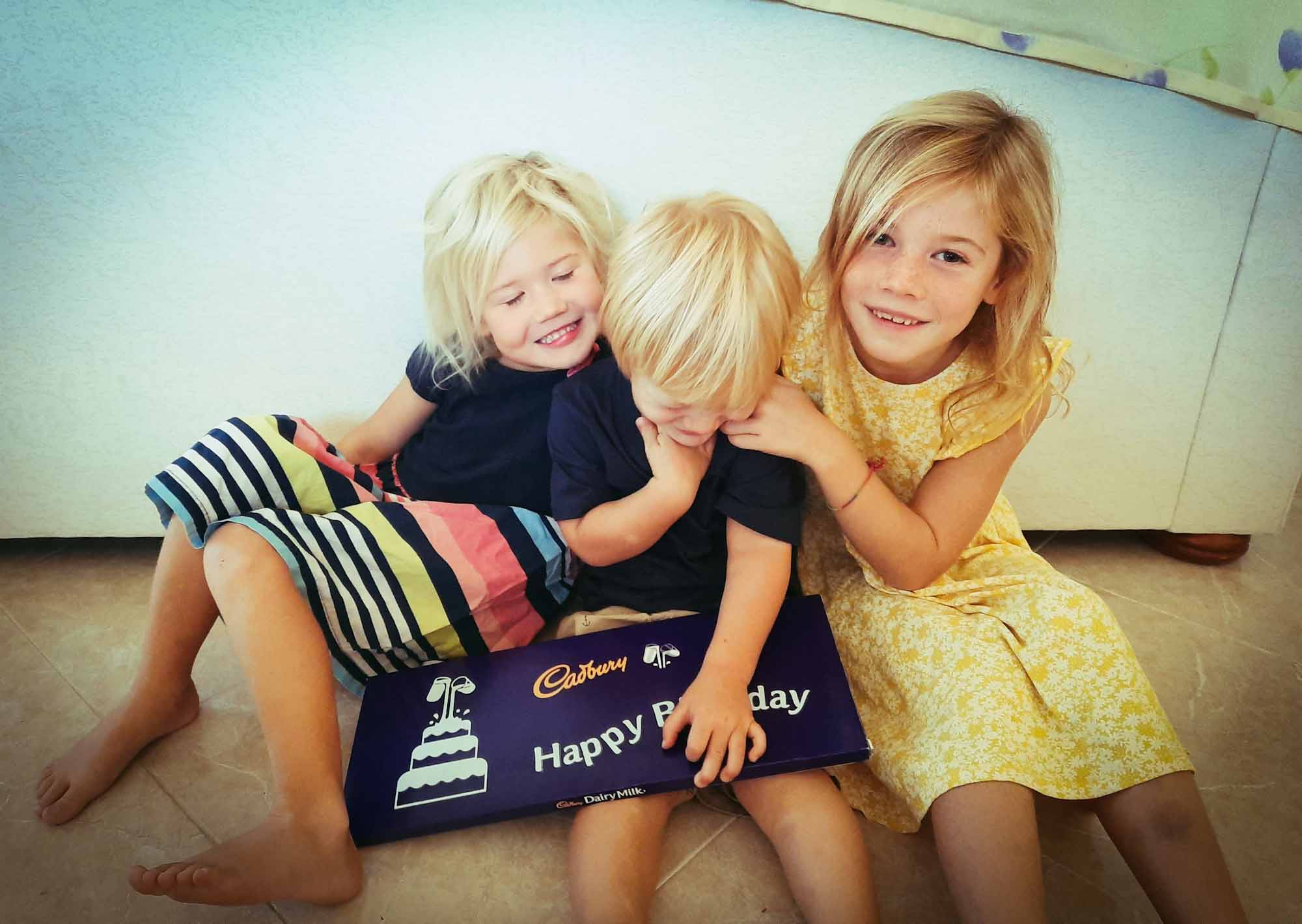 3 small children sat on the floor holding a giant chocolate bar with 'happy birthday' written on it