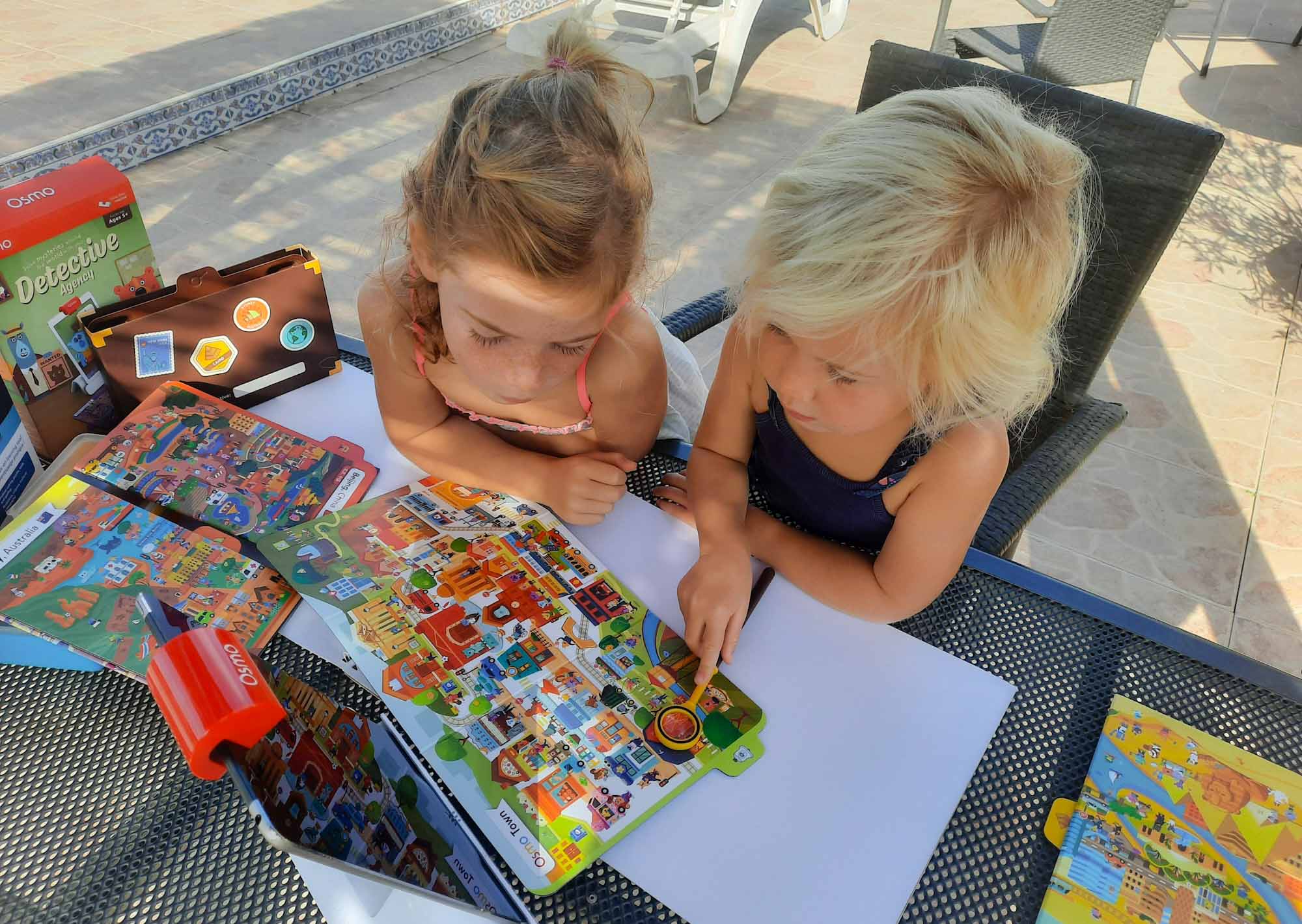 2 young girls with a toy magnifying glass, looking at a colourful cartoon map in front of an Ipod on a stand