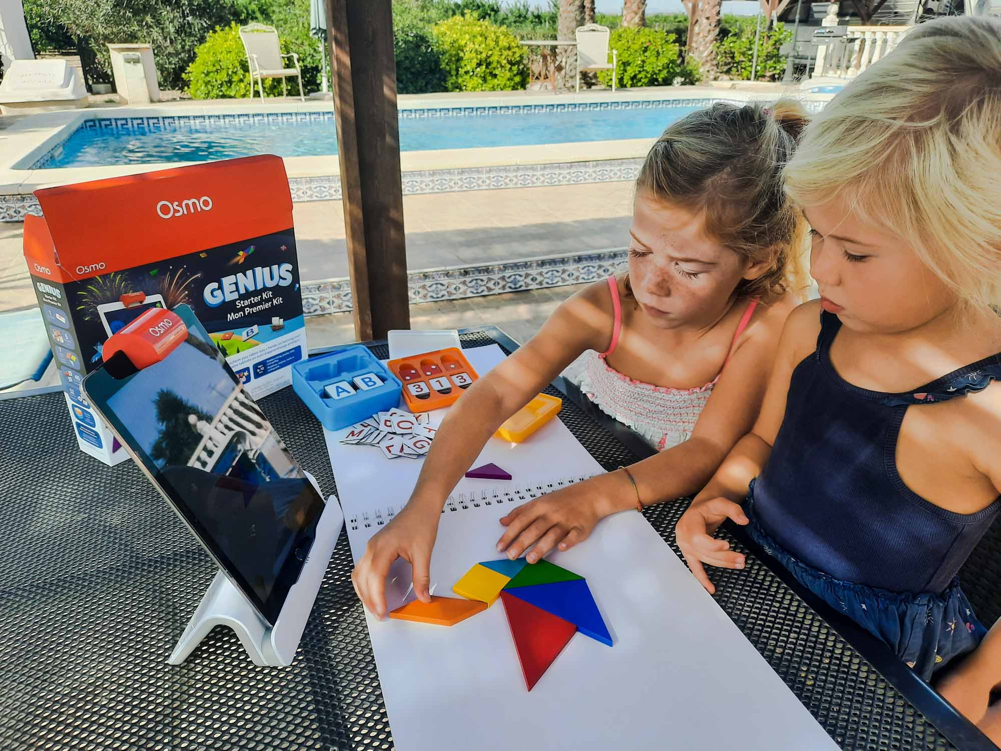 2 young girls making a picture on an outdoor table out of wooden shapes, while looking at an Ipad on a stand