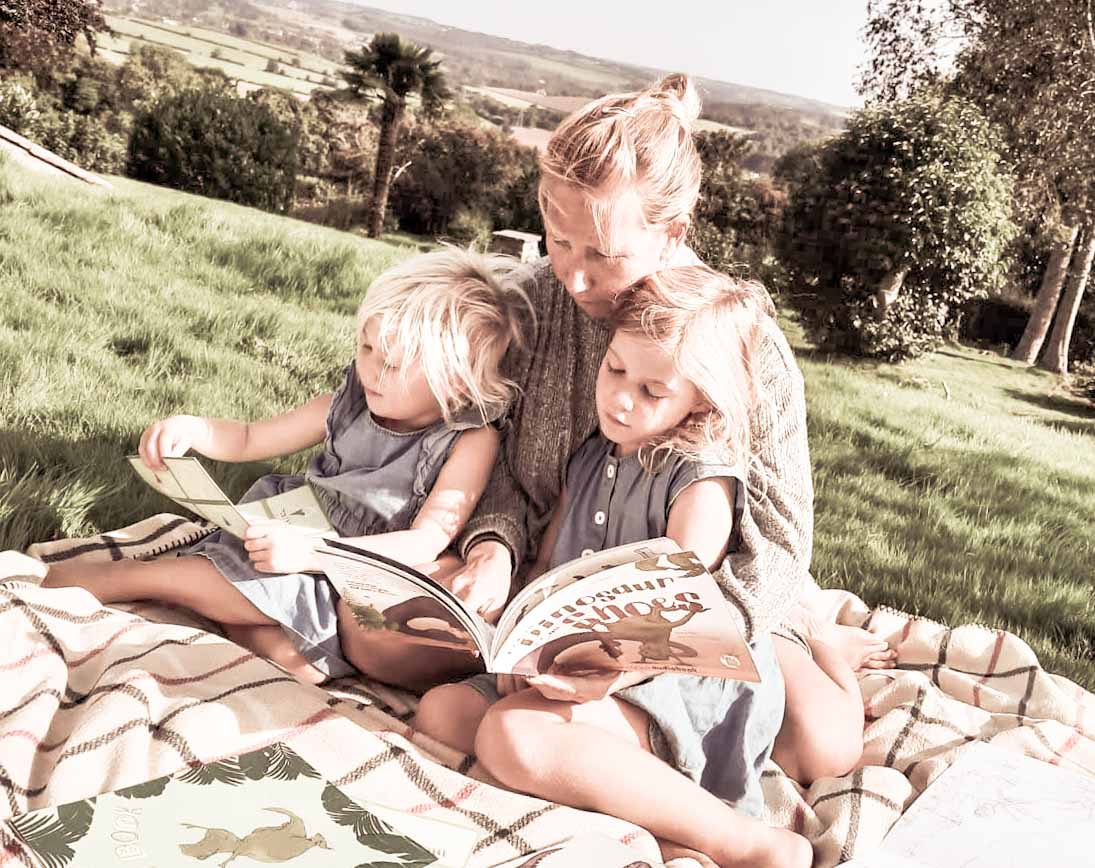 Mother and 2 daughters sat outside on a rug while reading a book together