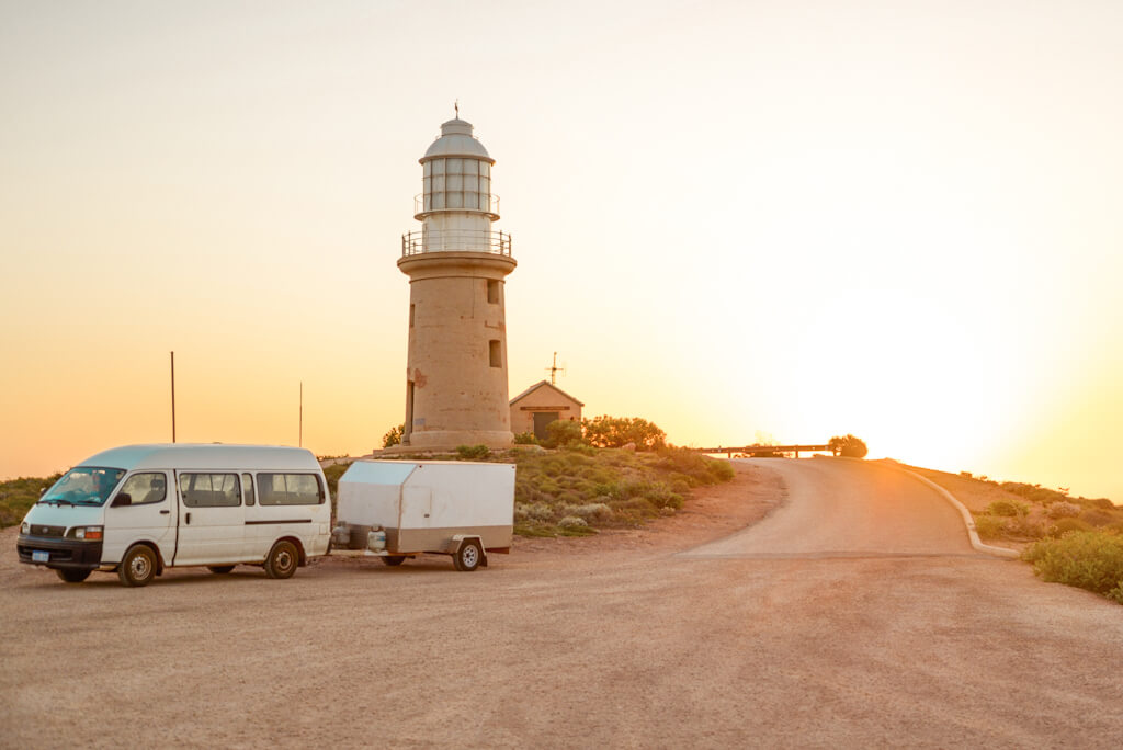 Campervan pulling a trailer, parked in front of a lighthouse at sunset