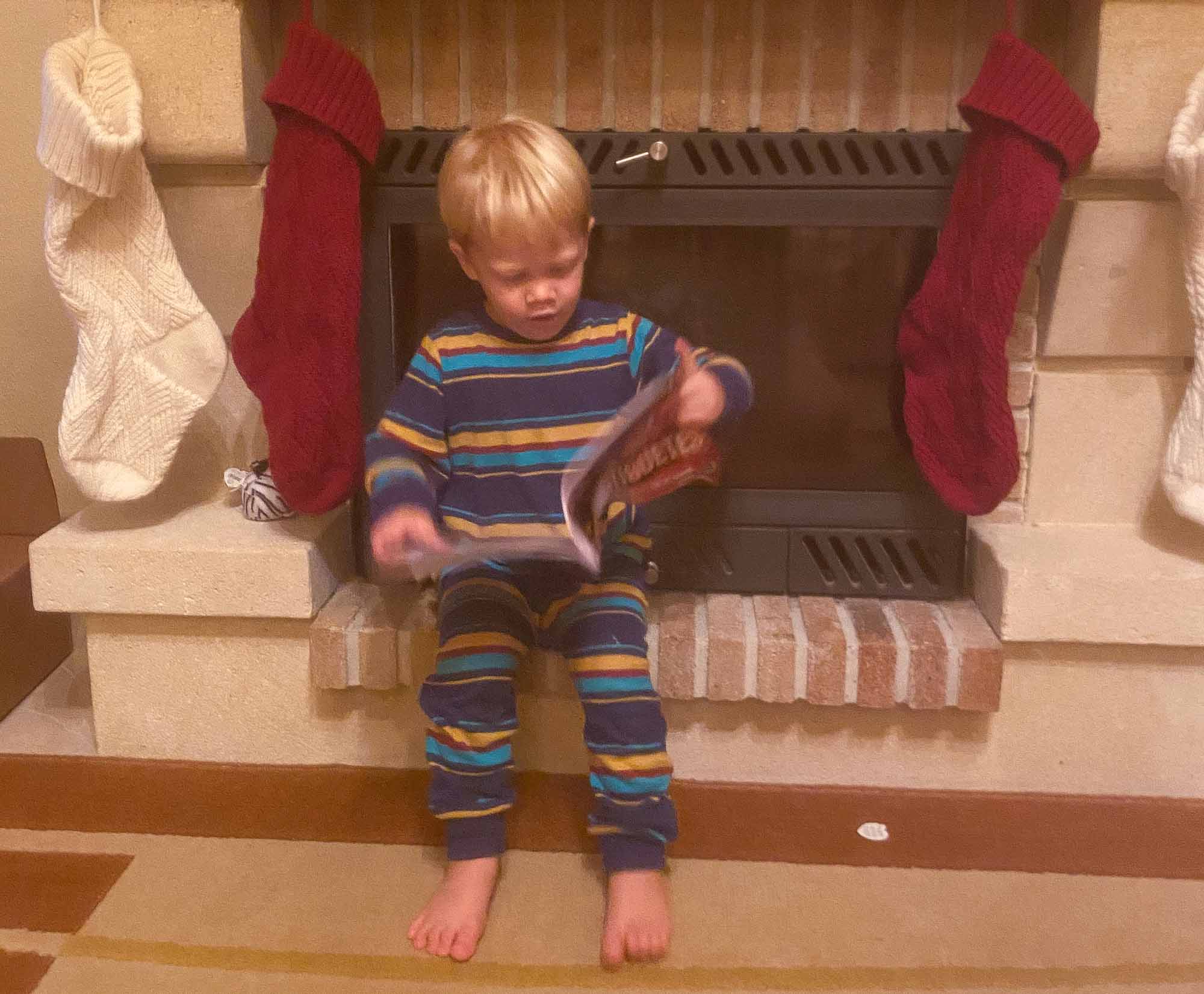Young boy wearing pyjamas while sat on a fireplace hearth, looking at a magazine