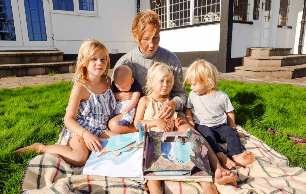 A family with 4 small children, sat outside on a rug while looking at a pop-up book