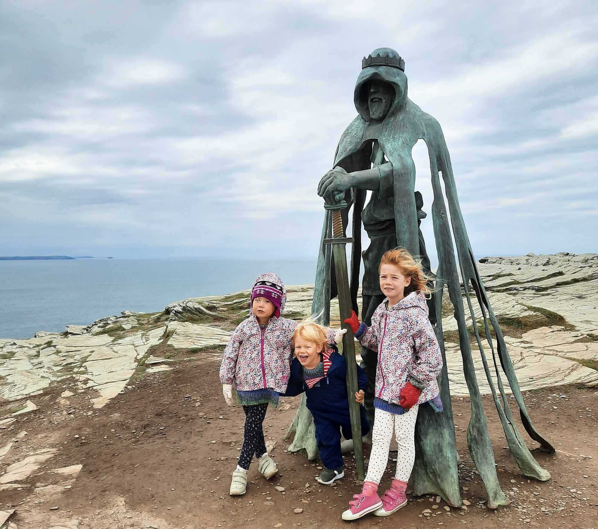 3 small children stood with a metal sculpture of King Arthur on a clifftop