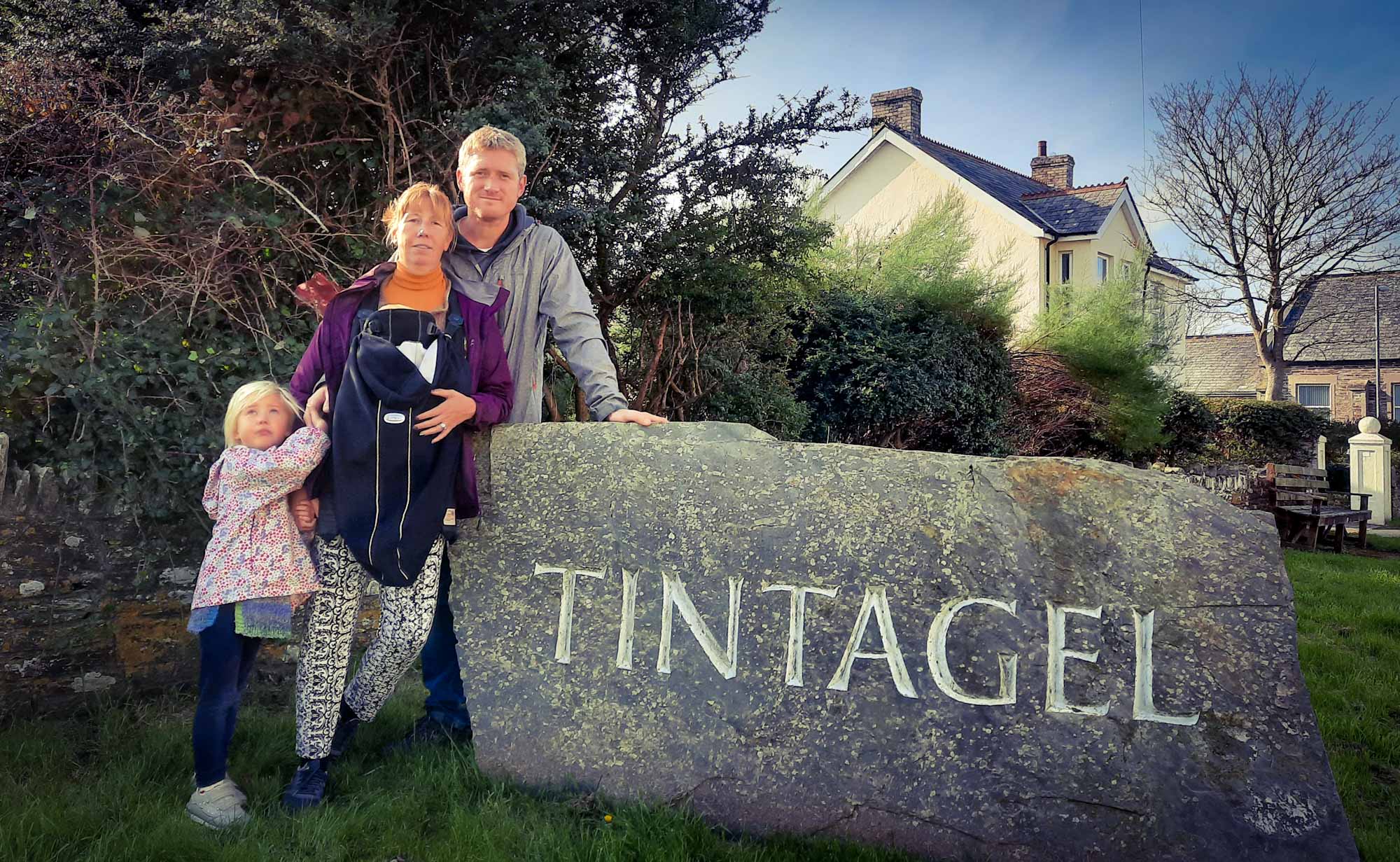 Family stood by a large, stone town sign, at the entrance to Tintagel, Cornwall, UK