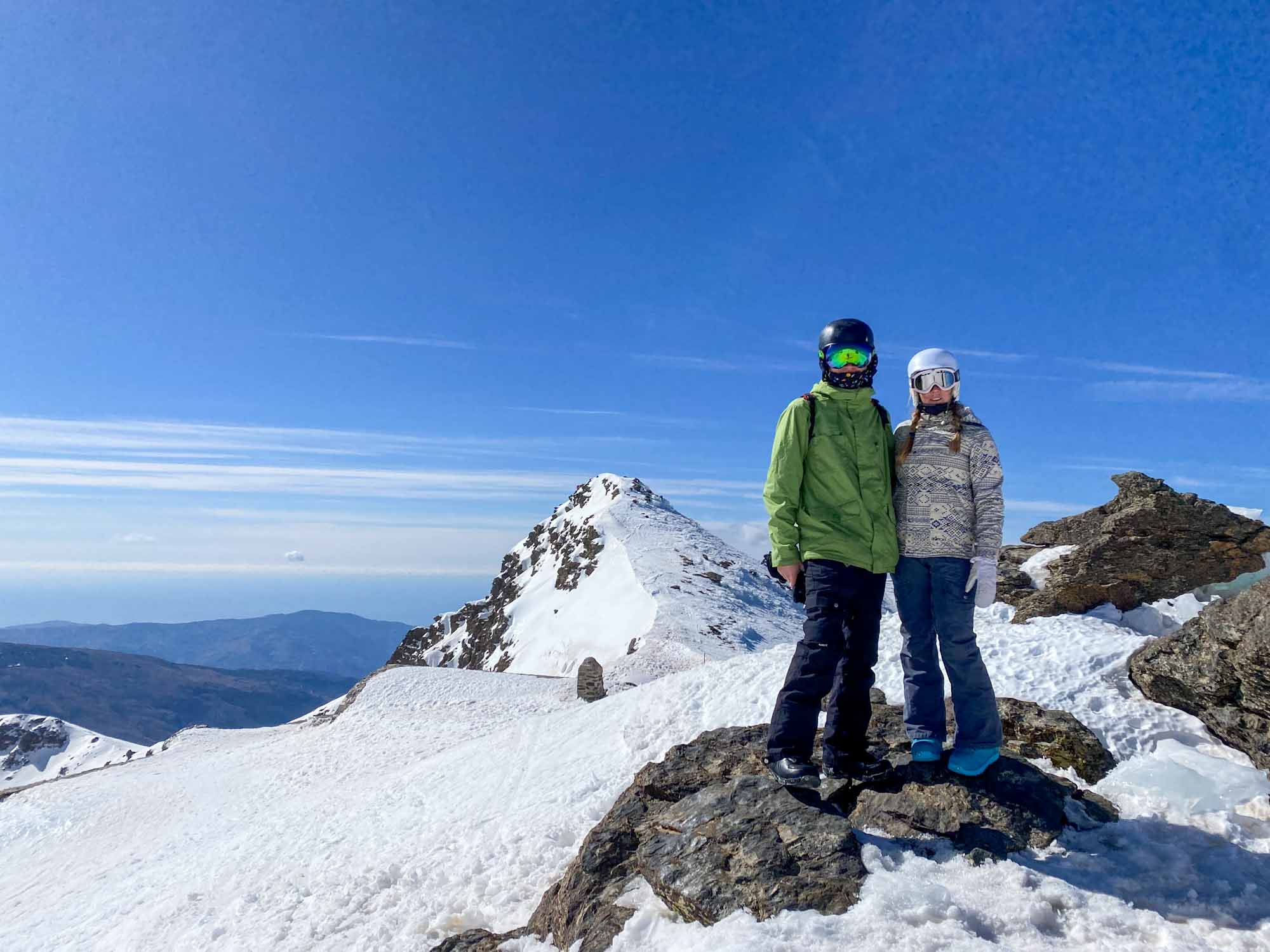 2 people wearing ski clothes stood on the snow on top of a mountain